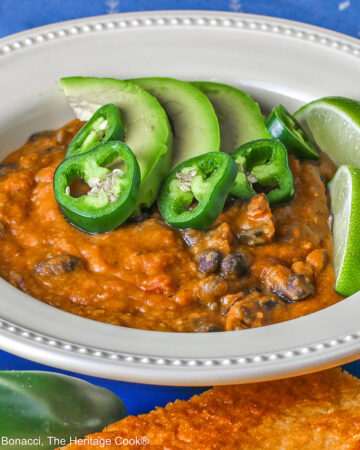 Pale yellow bowl of Vegan Red Lentil Chili with Pumpkin sitting on a blue plate surrounded by jalapeno peppers, cilantro, lime, and cornbread, with a blue cloth beneath © 2025 Jane Bonacci, The Heritage Cook.
