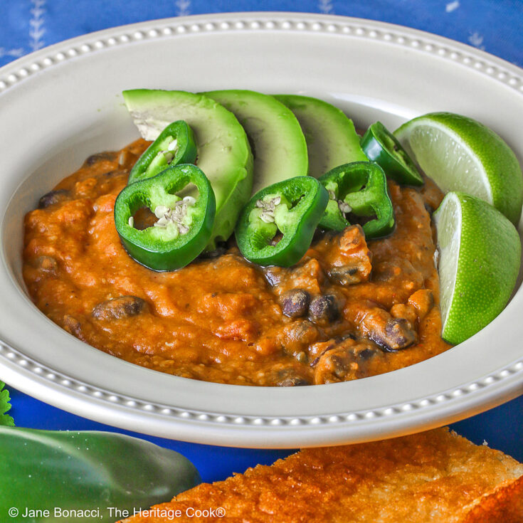 Pale yellow bowl of Vegan Red Lentil Chili with Pumpkin sitting on a blue plate surrounded by jalapeno peppers, cilantro, lime, and cornbread, with a blue cloth beneath © 2025 Jane Bonacci, The Heritage Cook.