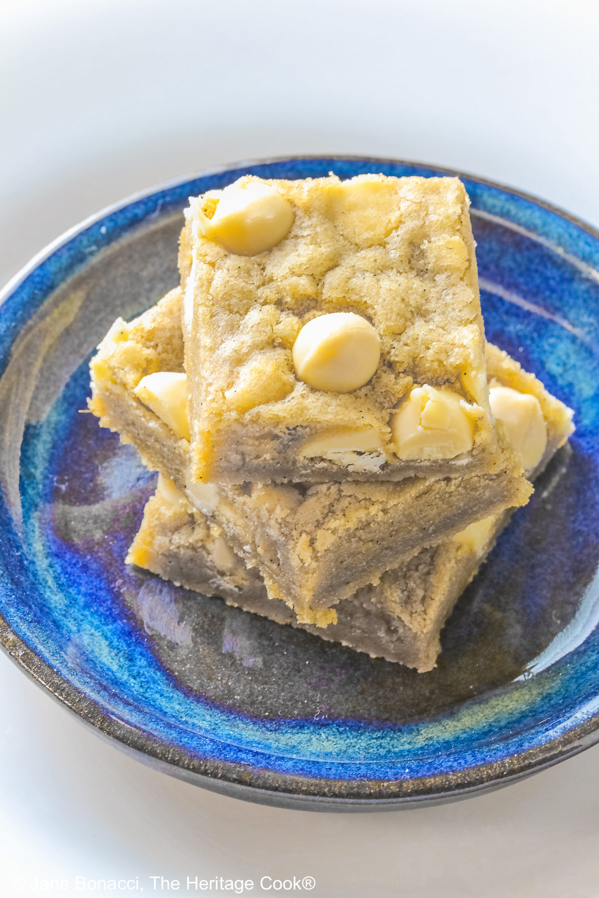 Small stack of Egg-Free White Chocolate Chip Blondies on a small dark blue plate, some on top of a little white plate, sitting on a red and white striped cloth on top of a mottled gray background © 2026 Jane Bonacci, The Heritage Cook.