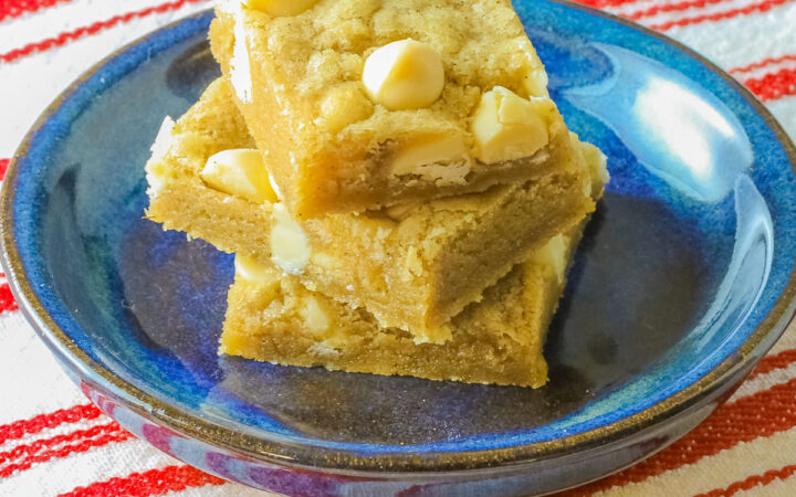 Small stack of Egg-Free White Chocolate Chip Blondies on a small dark blue plate, some on top of a little white plate, sitting on a red and white striped cloth on top of a mottled gray background © 2026 Jane Bonacci, The Heritage Cook.