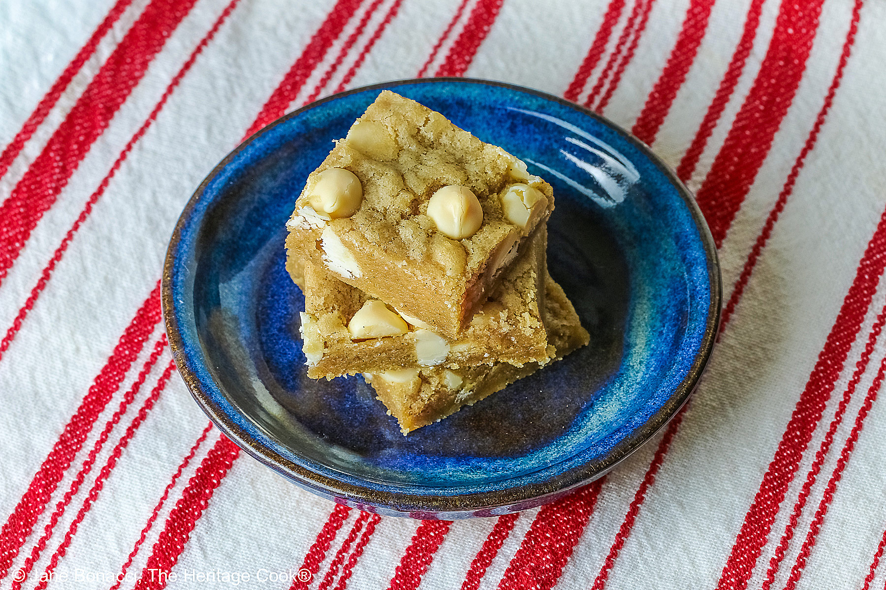 Small stack of Egg-Free White Chocolate Chip Blondies on a small dark blue plate, some on top of a little white plate, sitting on a red and white striped cloth on top of a mottled gray background © 2026 Jane Bonacci, The Heritage Cook.