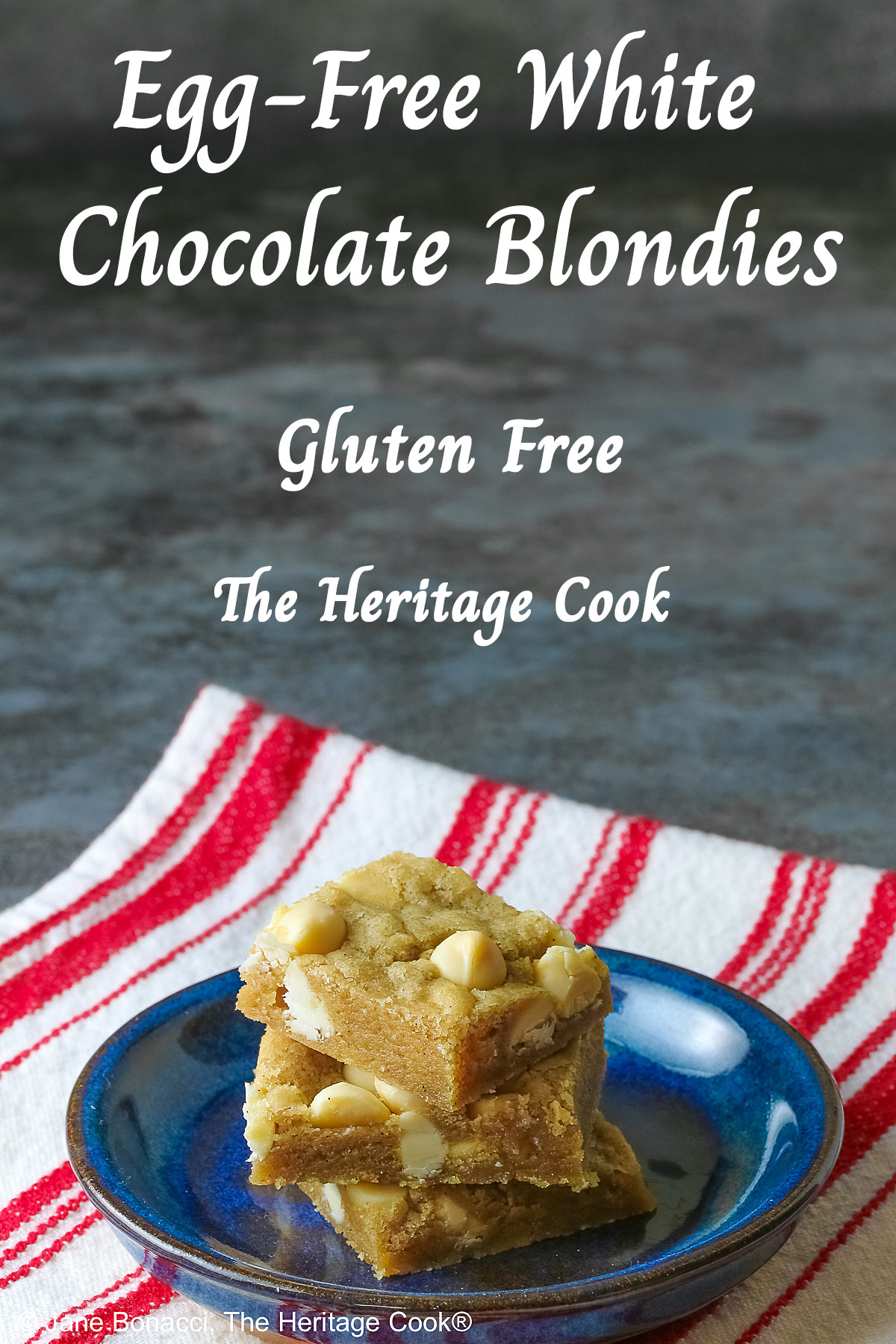 Small stack of Egg-Free White Chocolate Chip Blondies on a small dark blue plate, some on top of a little white plate, sitting on a red and white striped cloth on top of a mottled gray background © 2026 Jane Bonacci, The Heritage Cook.