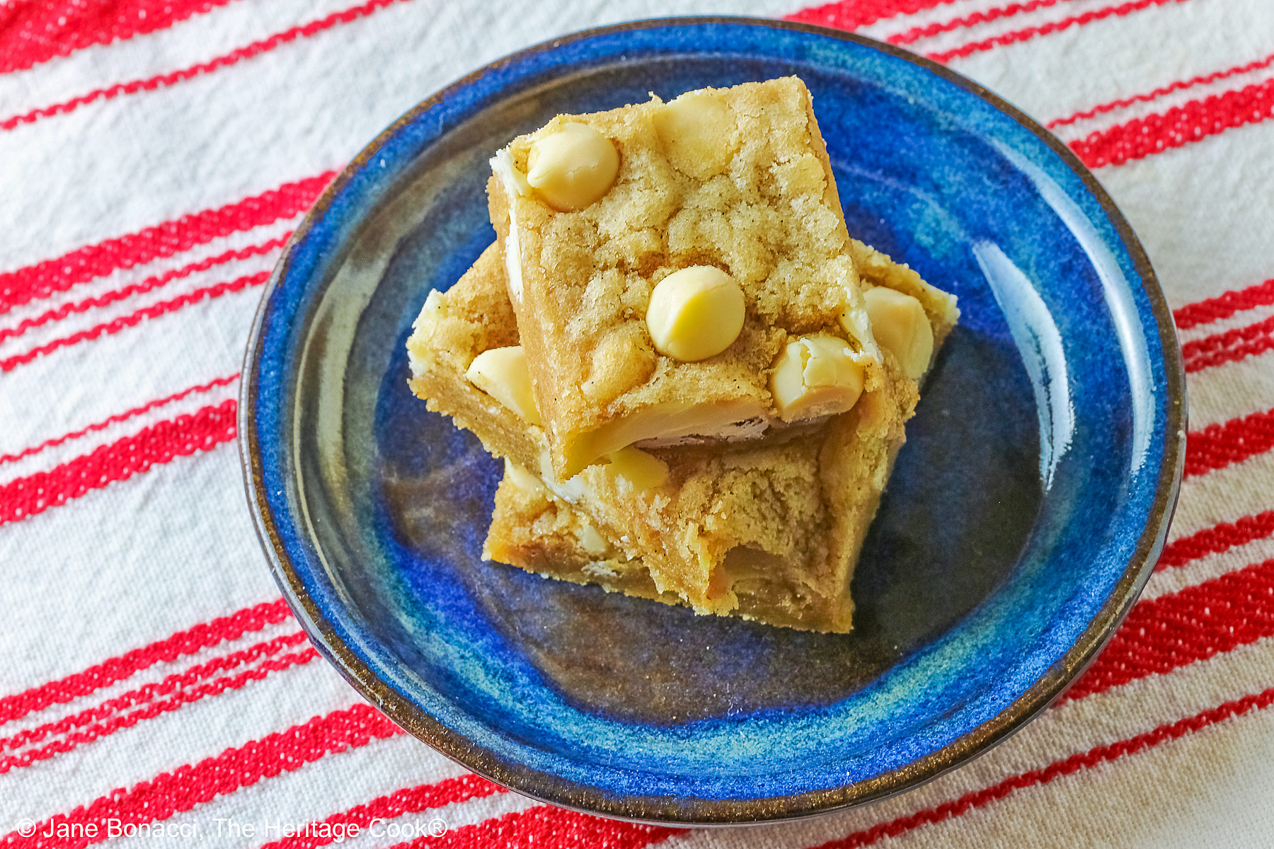 Small stack of Egg-Free White Chocolate Chip Blondies on a small dark blue plate, some on top of a little white plate, sitting on a red and white striped cloth on top of a mottled gray background © 2026 Jane Bonacci, The Heritage Cook.