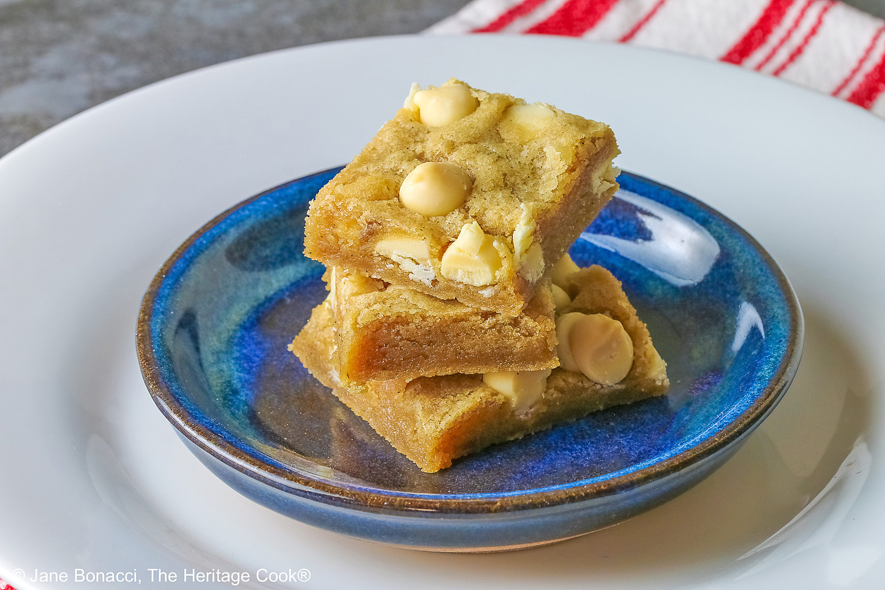 Small stack of Egg-Free White Chocolate Chip Blondies on a small dark blue plate, some on top of a little white plate, sitting on a red and white striped cloth on top of a mottled gray background © 2026 Jane Bonacci, The Heritage Cook.