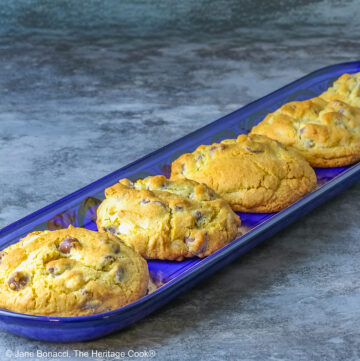 Five Massive Triple Chocolate Chip Cookies lined up on a long dark blue tray, sitting on a mottled blue-gray background; some with a cream cloth with forks on it and others with a blue and white checked cloth © 2026 Jane Bonacci, The Heritage Cook.