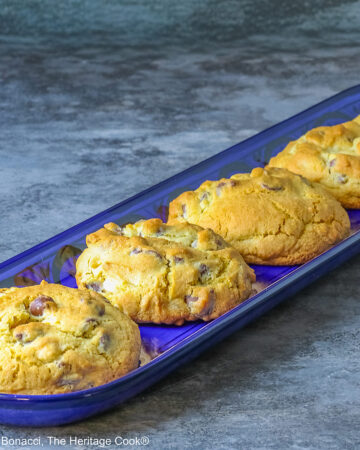 Five Massive Triple Chocolate Chip Cookies lined up on a long dark blue tray, sitting on a mottled blue-gray background; some with a cream cloth with forks on it and others with a blue and white checked cloth © 2026 Jane Bonacci, The Heritage Cook.