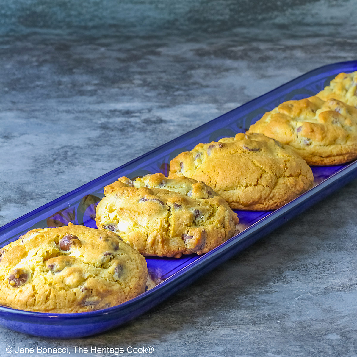 Five Massive Triple Chocolate Chip Cookies lined up on a long dark blue tray, sitting on a mottled blue-gray background; some with a cream cloth with forks on it and others with a blue and white checked cloth © 2026 Jane Bonacci, The Heritage Cook.