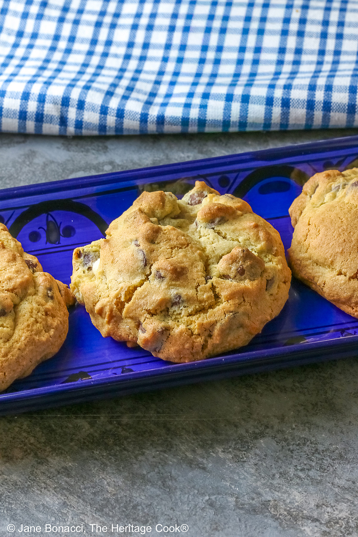 Five Massive Triple Chocolate Chip Cookies lined up on a long dark blue tray, sitting on a mottled blue-gray background; some with a cream cloth with forks on it and others with a blue and white checked cloth © 2026 Jane Bonacci, The Heritage Cook.