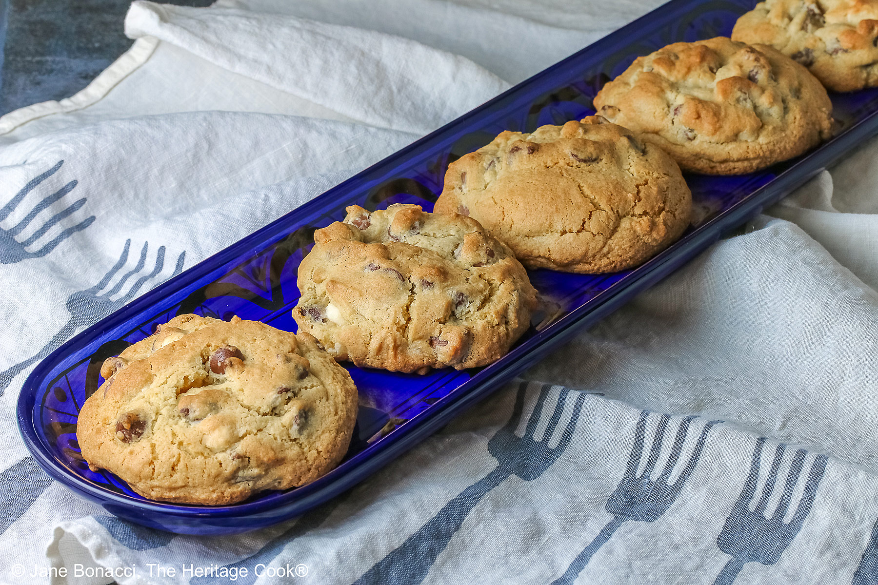 Five Massive Triple Chocolate Chip Cookies lined up on a long dark blue tray, sitting on a mottled blue-gray background; some with a cream cloth with forks on it and others with a blue and white checked cloth © 2026 Jane Bonacci, The Heritage Cook.