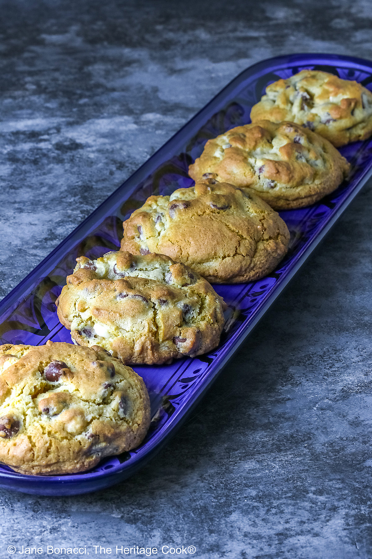 Five Massive Triple Chocolate Chip Cookies lined up on a long dark blue tray, sitting on a mottled blue-gray background; some with a cream cloth with forks on it and others with a blue and white checked cloth © 2026 Jane Bonacci, The Heritage Cook.