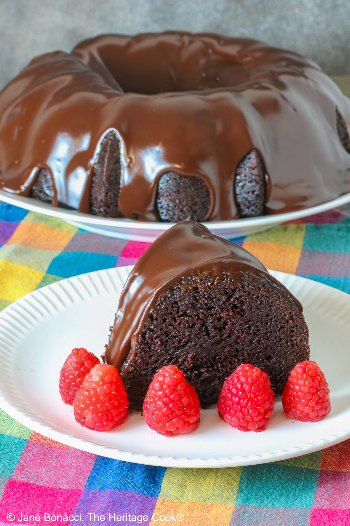 Chocolate Bourbon Bundt Cake on a white plate sitting on a brightly colored plaid cloth, some with a slice with a row of raspberries in front and a few with the whole cake behind © 2026 Jane Bonacci, The Heritage Cook.