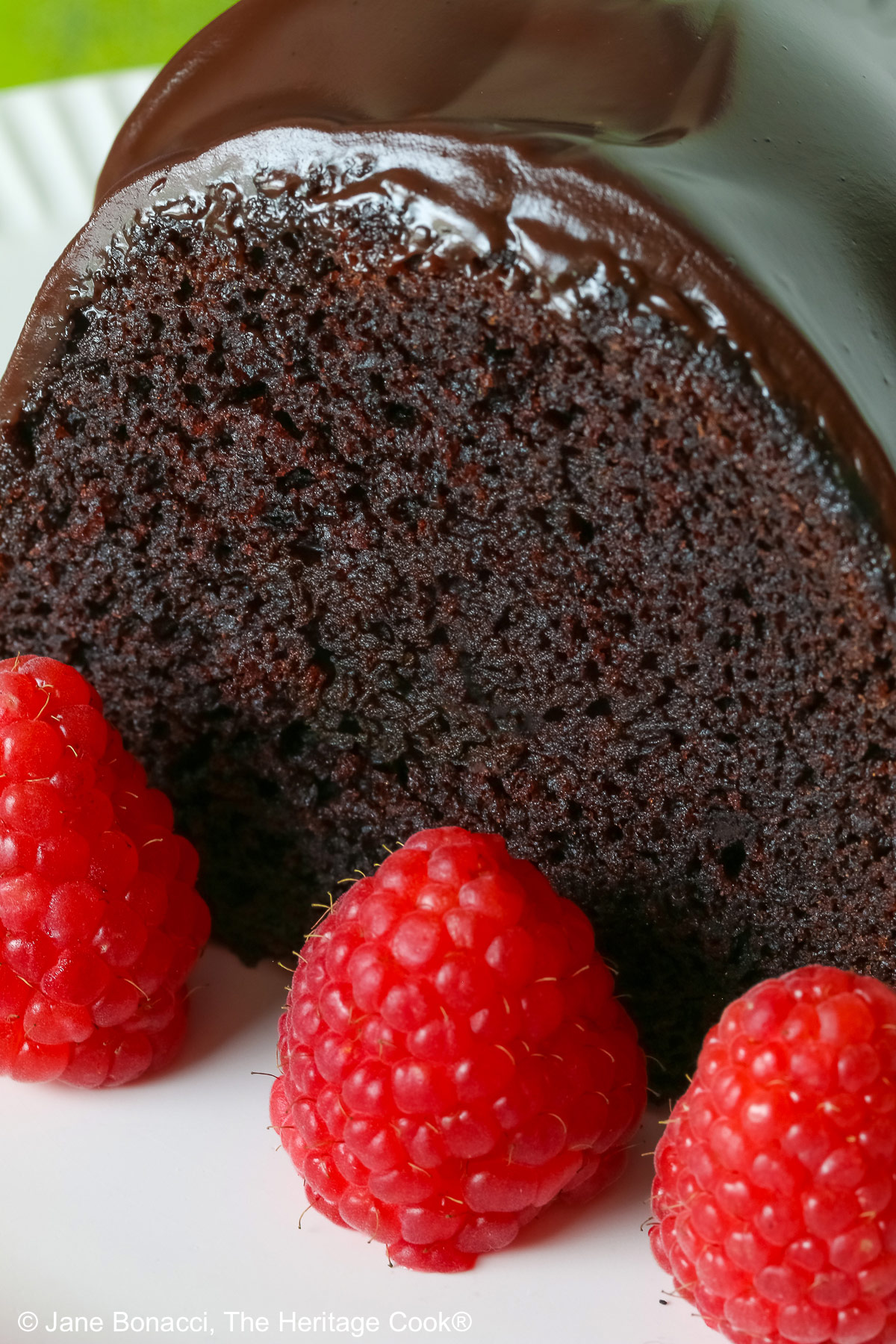 Chocolate Bourbon Bundt Cake on a white plate sitting on a brightly colored plaid cloth, some with a slice with a row of raspberries in front and a few with the whole cake behind © 2026 Jane Bonacci, The Heritage Cook.