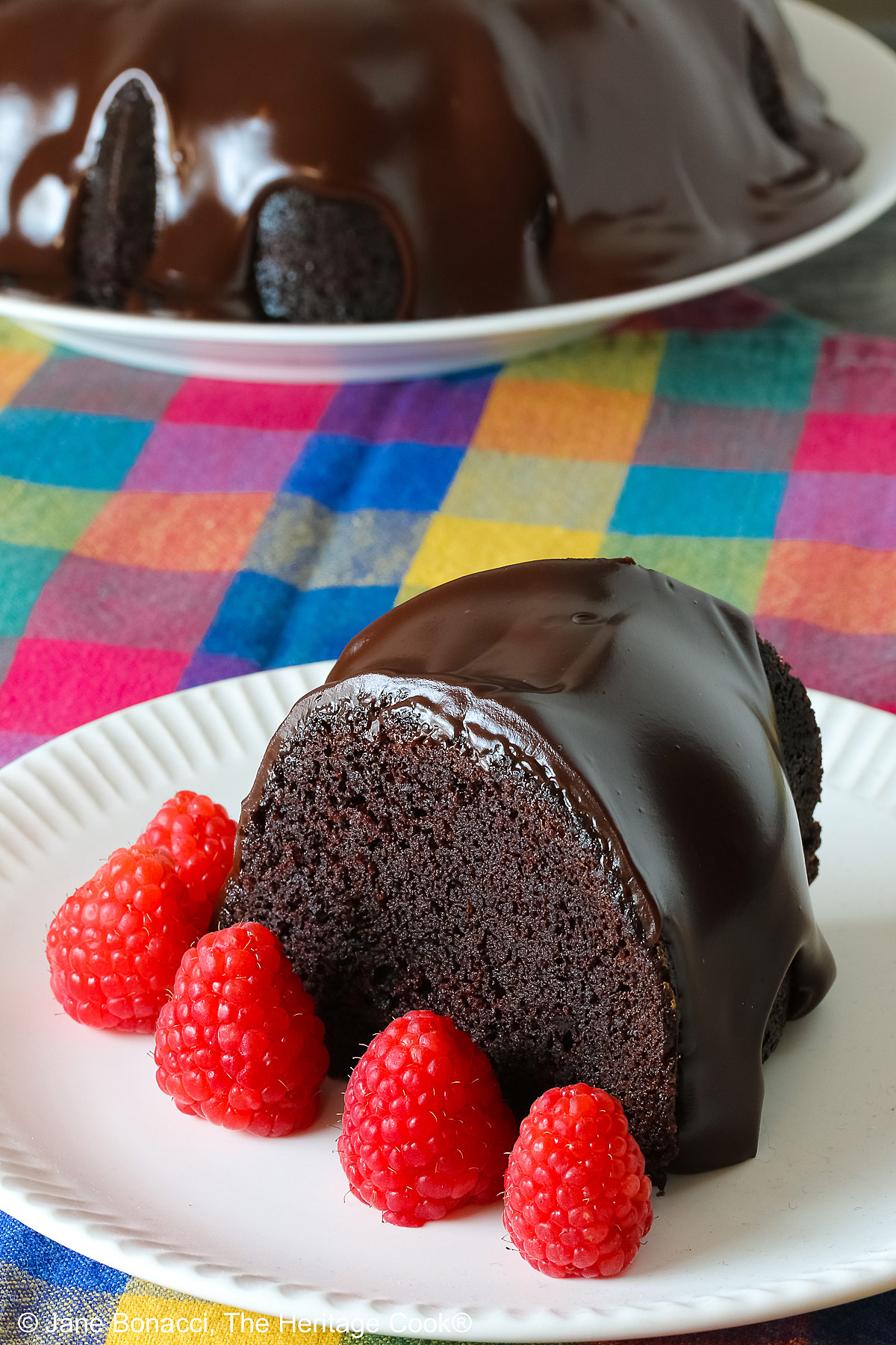 Chocolate Bourbon Bundt Cake on a white plate sitting on a brightly colored plaid cloth, some with a slice with a row of raspberries in front and a few with the whole cake behind © 2026 Jane Bonacci, The Heritage Cook.