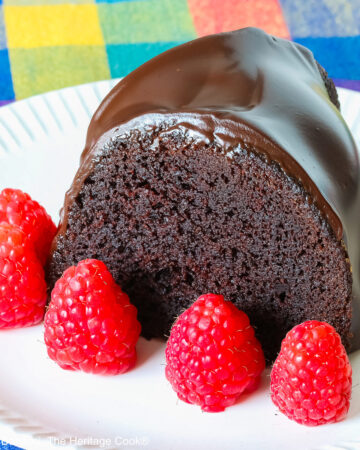 Chocolate Bourbon Bundt Cake on a white plate sitting on a brightly colored plaid cloth, some with a slice with a row of raspberries in front and a few with the whole cake behind © 2026 Jane Bonacci, The Heritage Cook.