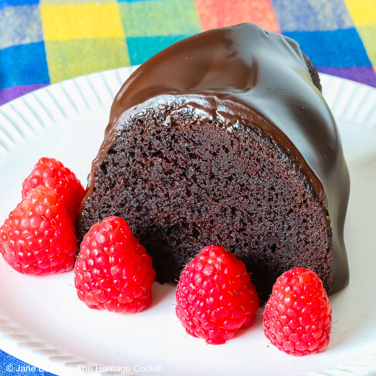 Chocolate Bourbon Bundt Cake on a white plate sitting on a brightly colored plaid cloth, some with a slice with a row of raspberries in front and a few with the whole cake behind © 2026 Jane Bonacci, The Heritage Cook.