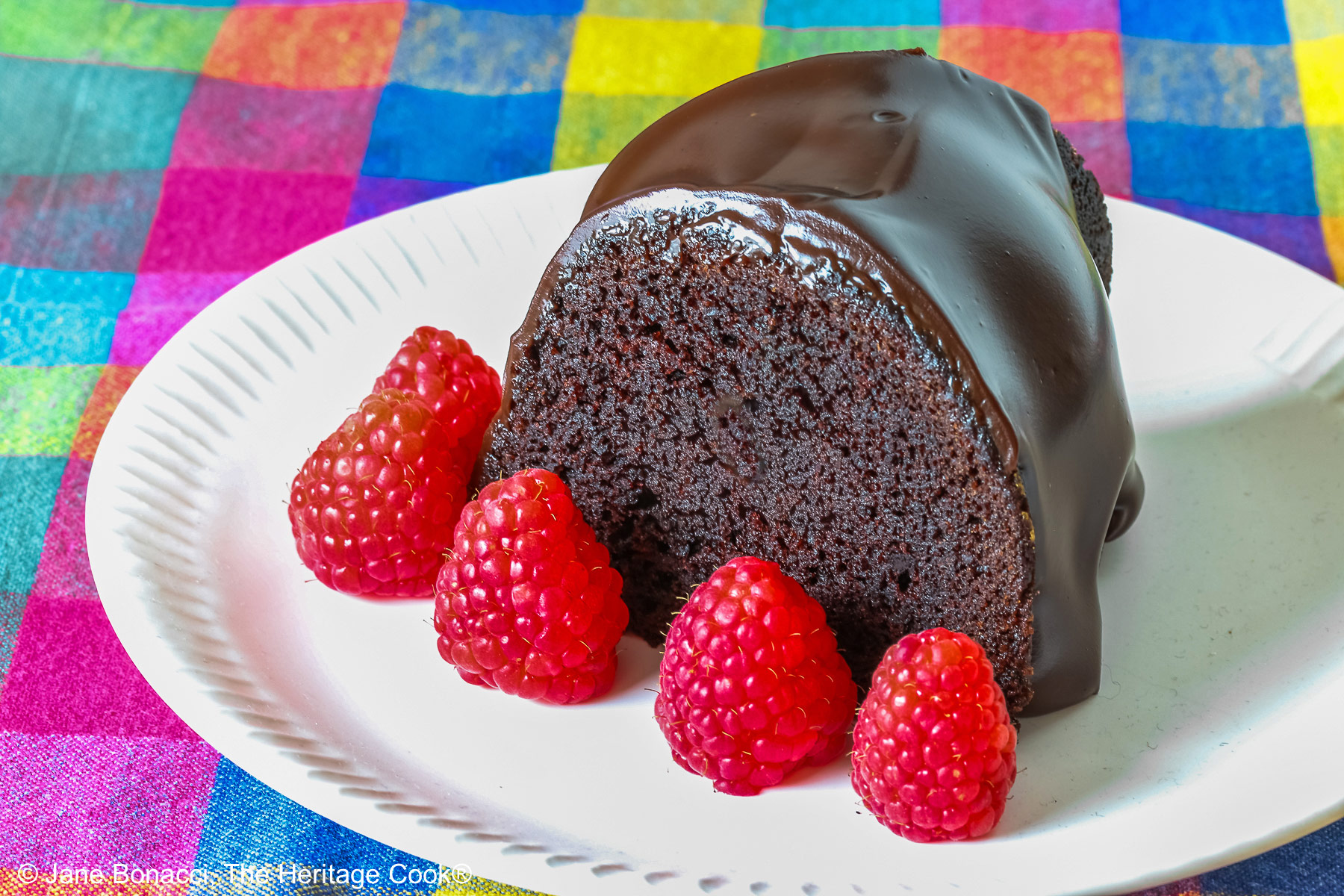 Chocolate Bourbon Bundt Cake on a white plate sitting on a brightly colored plaid cloth, some with a slice with a row of raspberries in front and a few with the whole cake behind © 2026 Jane Bonacci, The Heritage Cook.