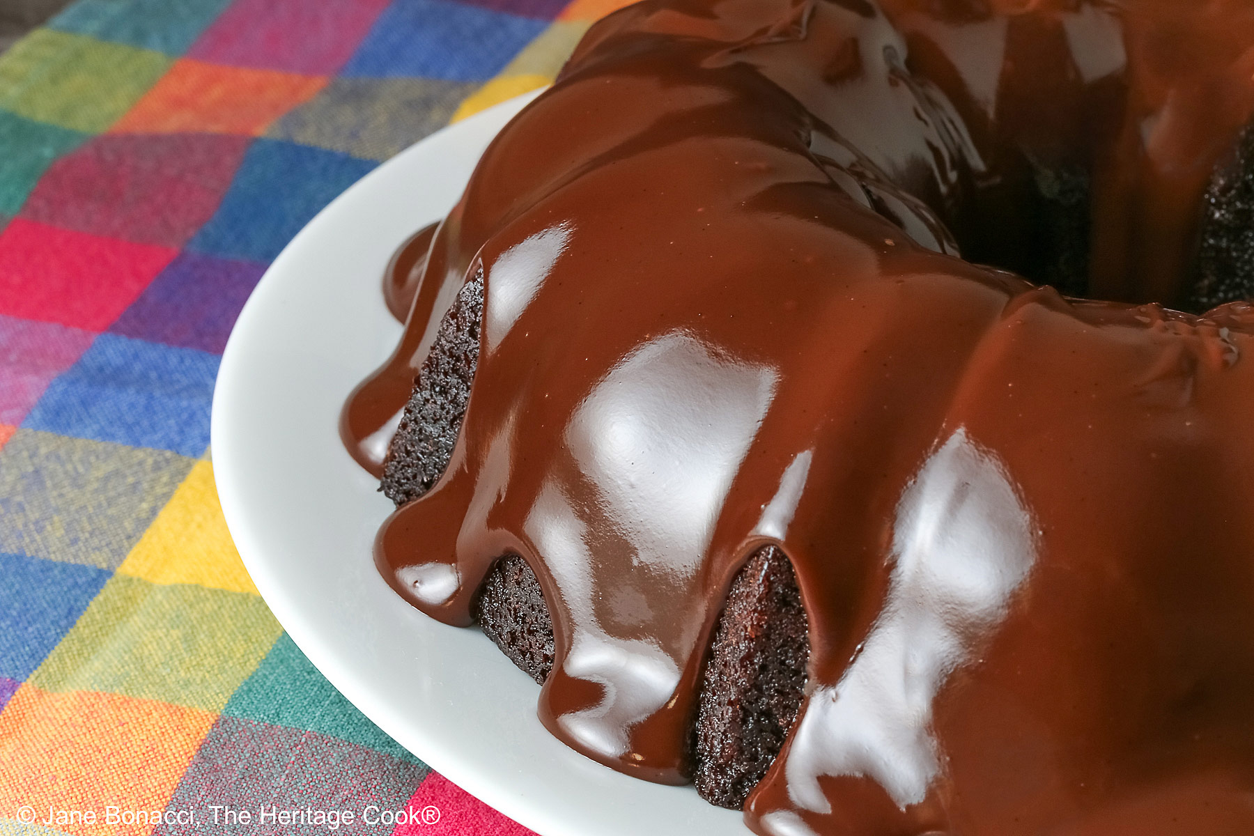 Chocolate Bourbon Bundt Cake on a white plate sitting on a brightly colored plaid cloth, some with a slice with a row of raspberries in front and a few with the whole cake behind © 2026 Jane Bonacci, The Heritage Cook.