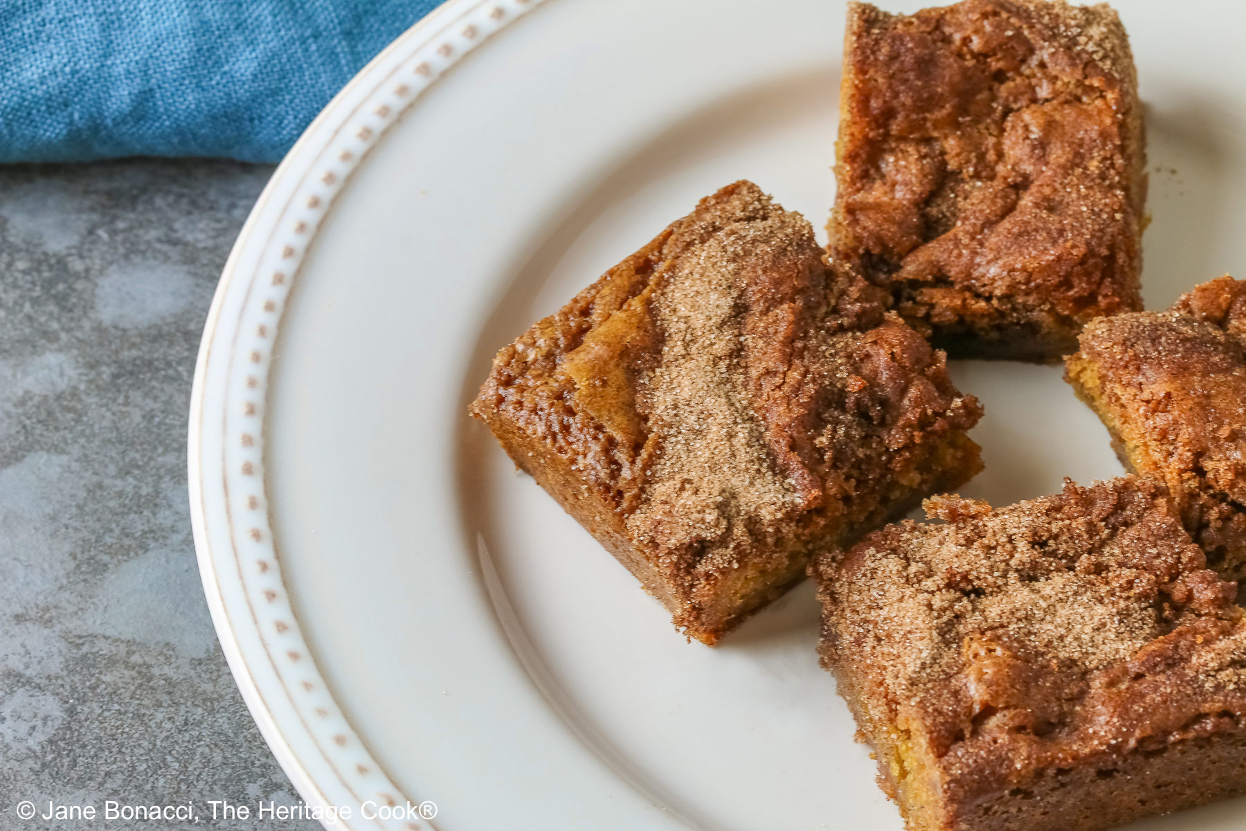 Four Cinnamon Sugar Blondies on a creamy white plate with small beads around the rim, set in a circle and stacks of two each, with a pale blue-teal cloth on a mottled gray background © 2026 Jane Bonacci, The Heritage Cook.