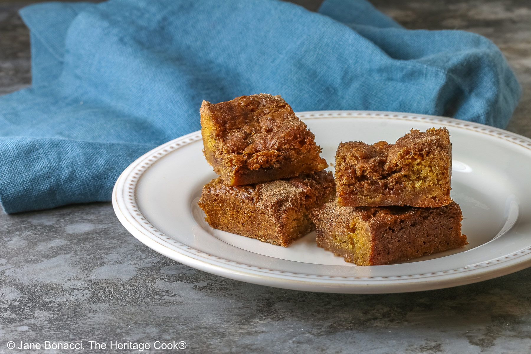 Four Cinnamon Sugar Blondies on a creamy white plate with small beads around the rim, set in a circle and stacks of two each, with a pale blue-teal cloth on a mottled gray background © 2026 Jane Bonacci, The Heritage Cook.