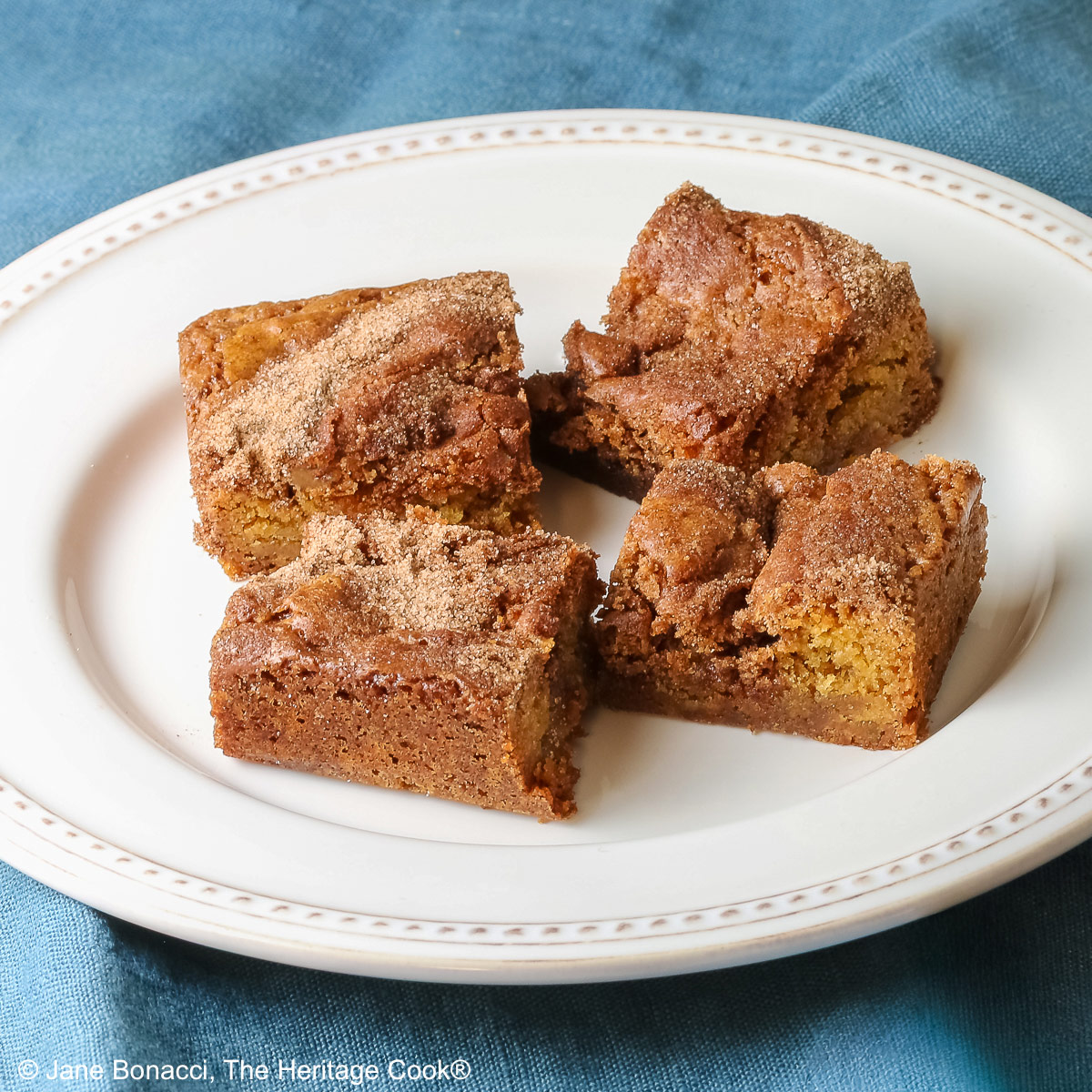 Four Cinnamon Sugar Blondies on a creamy white plate with small beads around the rim, set in a circle and stacks of two each, with a pale blue-teal cloth on a mottled gray background © 2026 Jane Bonacci, The Heritage Cook.