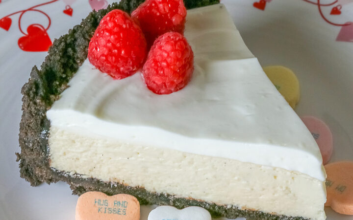 A single slice of Egg Free Cheesecake with Oreo Crust on a plate decorated with red hearts and flowers, sitting on a red and white striped cloth, with candy Valentine’s hearts around the edges and 3 bright red raspberries on the top © 2026 Jane Bonacci, The Heritage Cook.