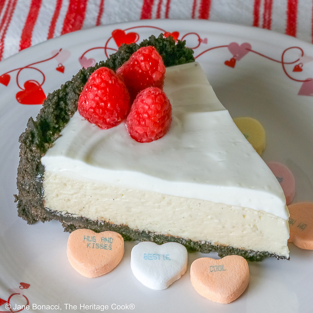 A single slice of Egg Free Cheesecake with Oreo Crust on a plate decorated with red hearts and flowers, sitting on a red and white striped cloth, with candy Valentine’s hearts around the edges and 3 bright red raspberries on the top © 2026 Jane Bonacci, The Heritage Cook.