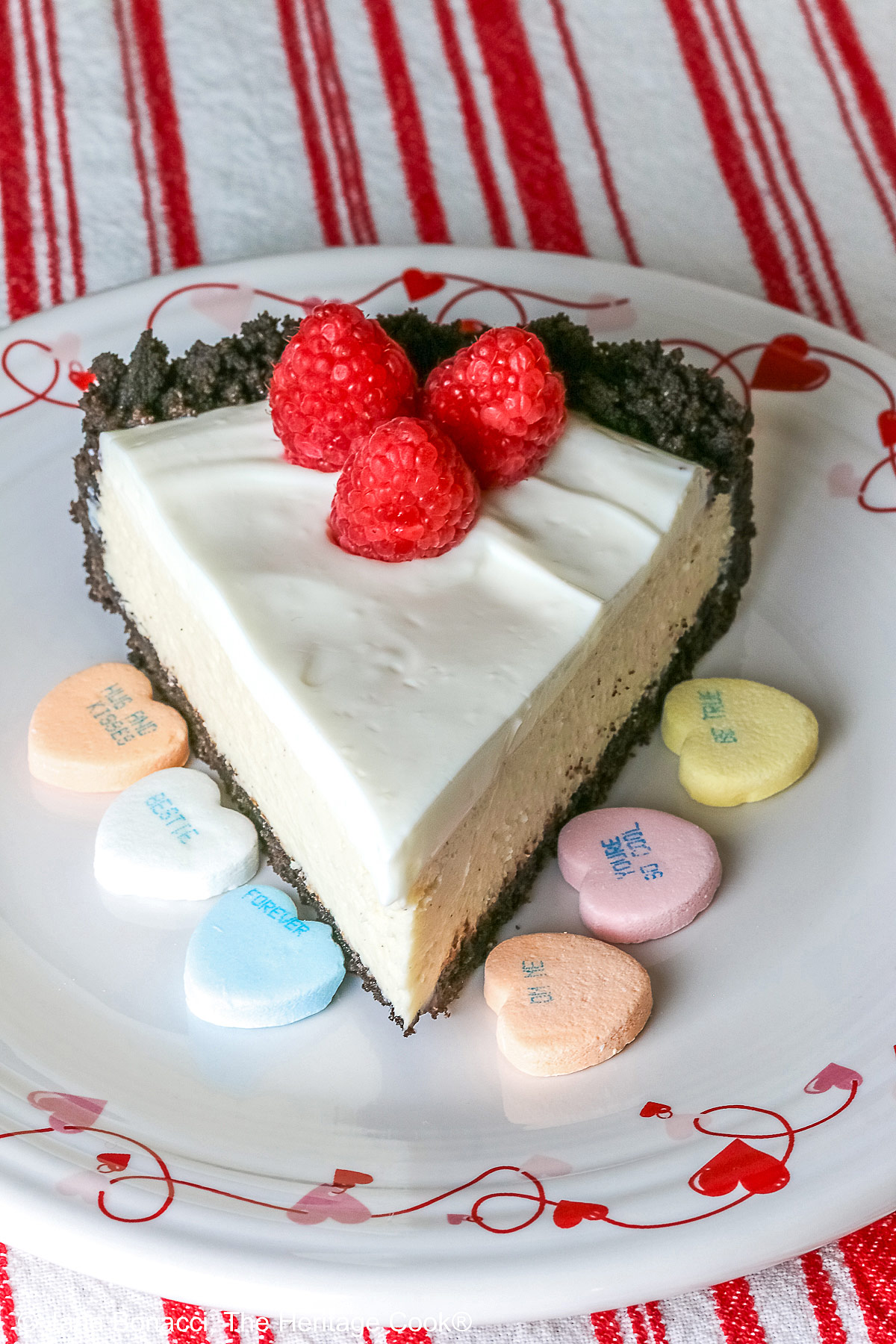 A single slice of Egg Free Cheesecake with Oreo Crust on a plate decorated with red hearts and flowers, sitting on a red and white striped cloth, with candy Valentine’s hearts around the edges and 3 bright red raspberries on the top © 2026 Jane Bonacci, The Heritage Cook.