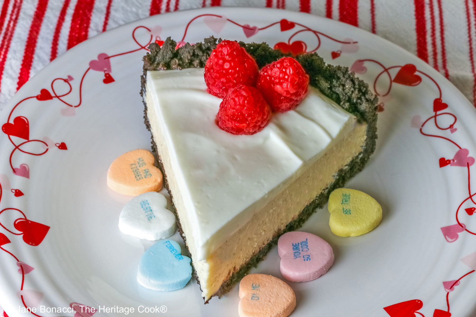A single slice of Egg Free Cheesecake with Oreo Crust on a plate decorated with red hearts and flowers, sitting on a red and white striped cloth, with candy Valentine’s hearts around the edges and 3 bright red raspberries on the top © 2026 Jane Bonacci, The Heritage Cook.