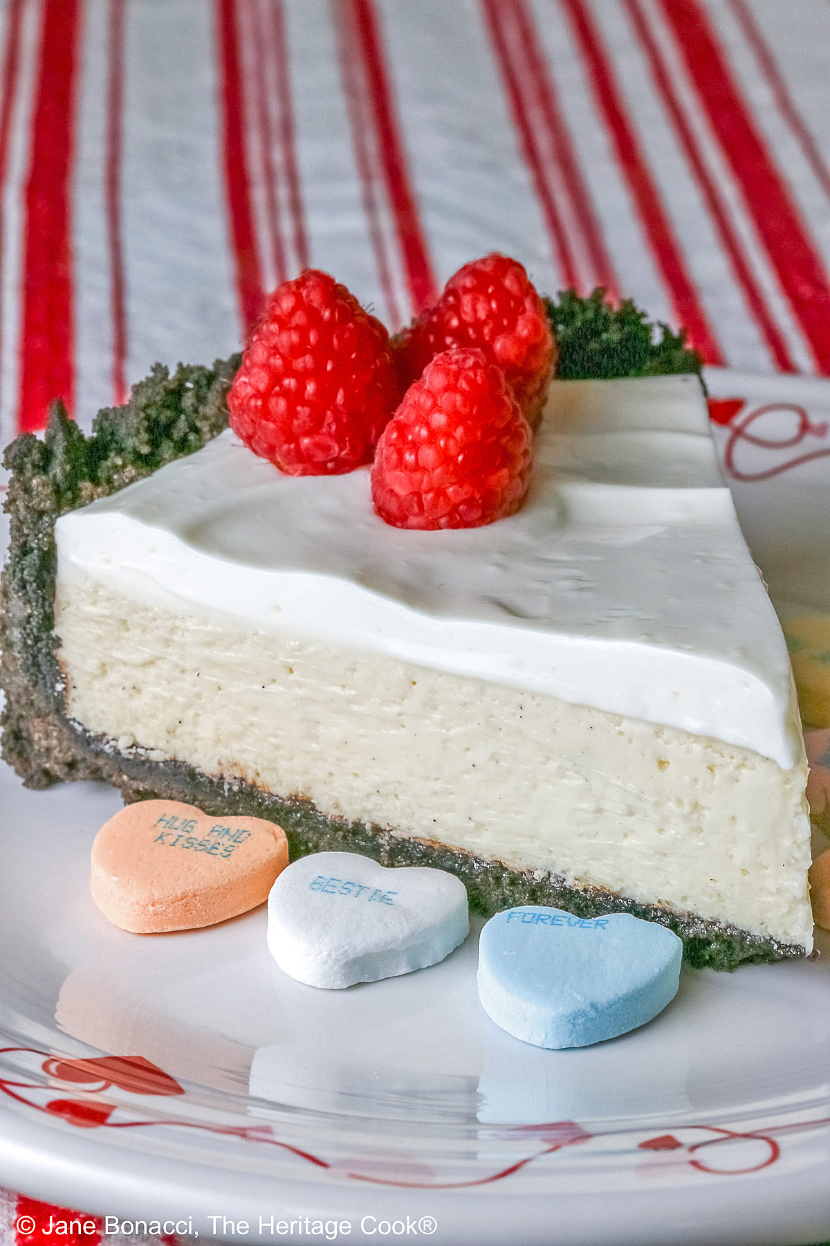 A single slice of Egg Free Cheesecake with Oreo Crust on a plate decorated with red hearts and flowers, sitting on a red and white striped cloth, with candy Valentine’s hearts around the edges and 3 bright red raspberries on the top © 2026 Jane Bonacci, The Heritage Cook.