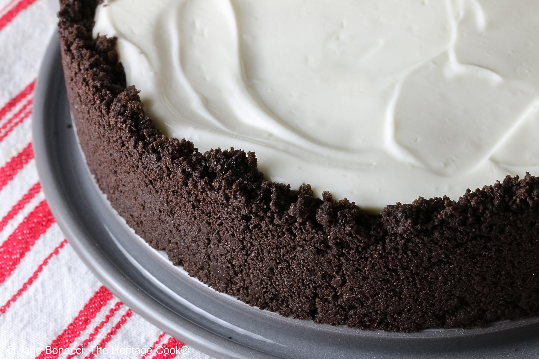 A single slice of Egg Free Cheesecake with Oreo Crust on a plate decorated with red hearts and flowers, sitting on a red and white striped cloth, with candy Valentine’s hearts around the edges and 3 bright red raspberries on the top © 2026 Jane Bonacci, The Heritage Cook.
