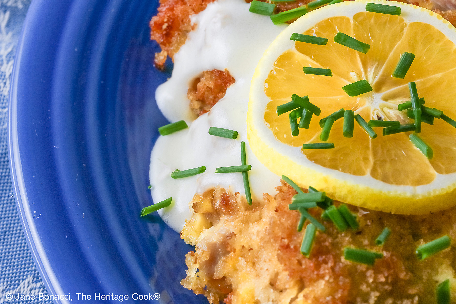 A golden brown crisp fried chicken thigh with a lemon-cream-garlic-butter sauce over the center, topped with a lemon slice and chopped chives, flanked with yellow saffron rice and green broccolini on a blue plate with a blue cloth beneath © 2026 Jane Bonacci, The Heritage Cook.