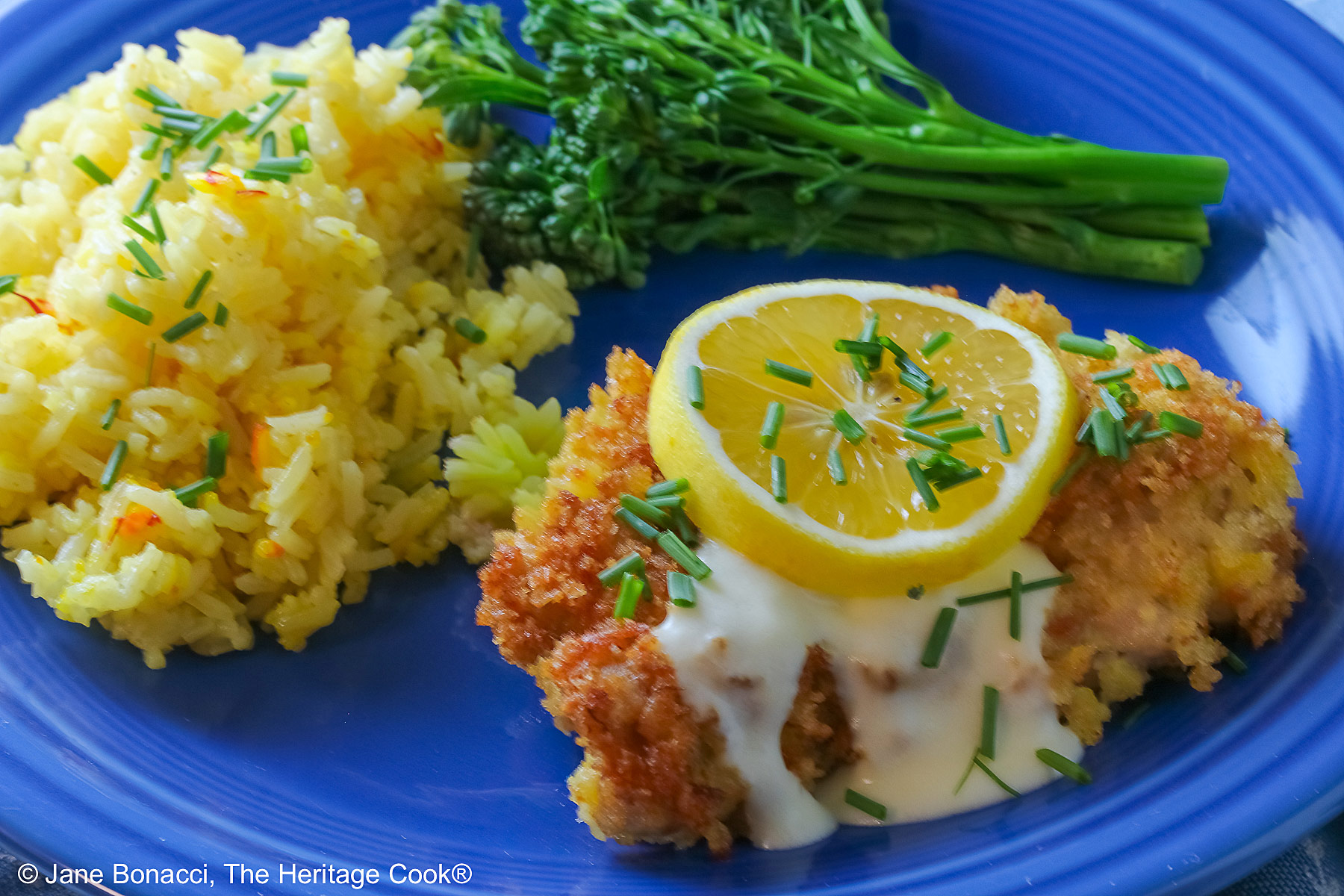 A golden brown crisp fried chicken thigh with a lemon-cream-garlic-butter sauce over the center, topped with a lemon slice and chopped chives, flanked with yellow saffron rice and green broccolini on a blue plate with a blue cloth beneath © 2026 Jane Bonacci, The Heritage Cook.
