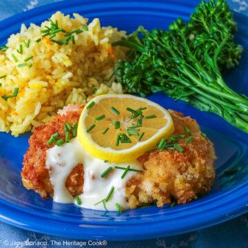 A golden brown crisp fried chicken thigh with a lemon-cream-garlic-butter sauce over the center, topped with a lemon slice and chopped chives, flanked with yellow saffron rice and green broccolini on a blue plate with a blue cloth beneath © 2026 Jane Bonacci, The Heritage Cook.