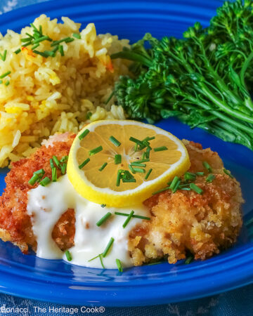 A golden brown crisp fried chicken thigh with a lemon-cream-garlic-butter sauce over the center, topped with a lemon slice and chopped chives, flanked with yellow saffron rice and green broccolini on a blue plate with a blue cloth beneath © 2026 Jane Bonacci, The Heritage Cook.