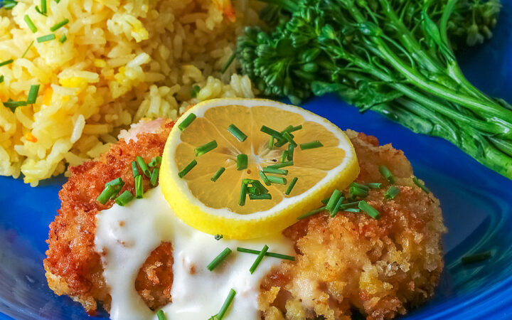 A golden brown crisp fried chicken thigh with a lemon-cream-garlic-butter sauce over the center, topped with a lemon slice and chopped chives, flanked with yellow saffron rice and green broccolini on a blue plate with a blue cloth beneath © 2026 Jane Bonacci, The Heritage Cook.