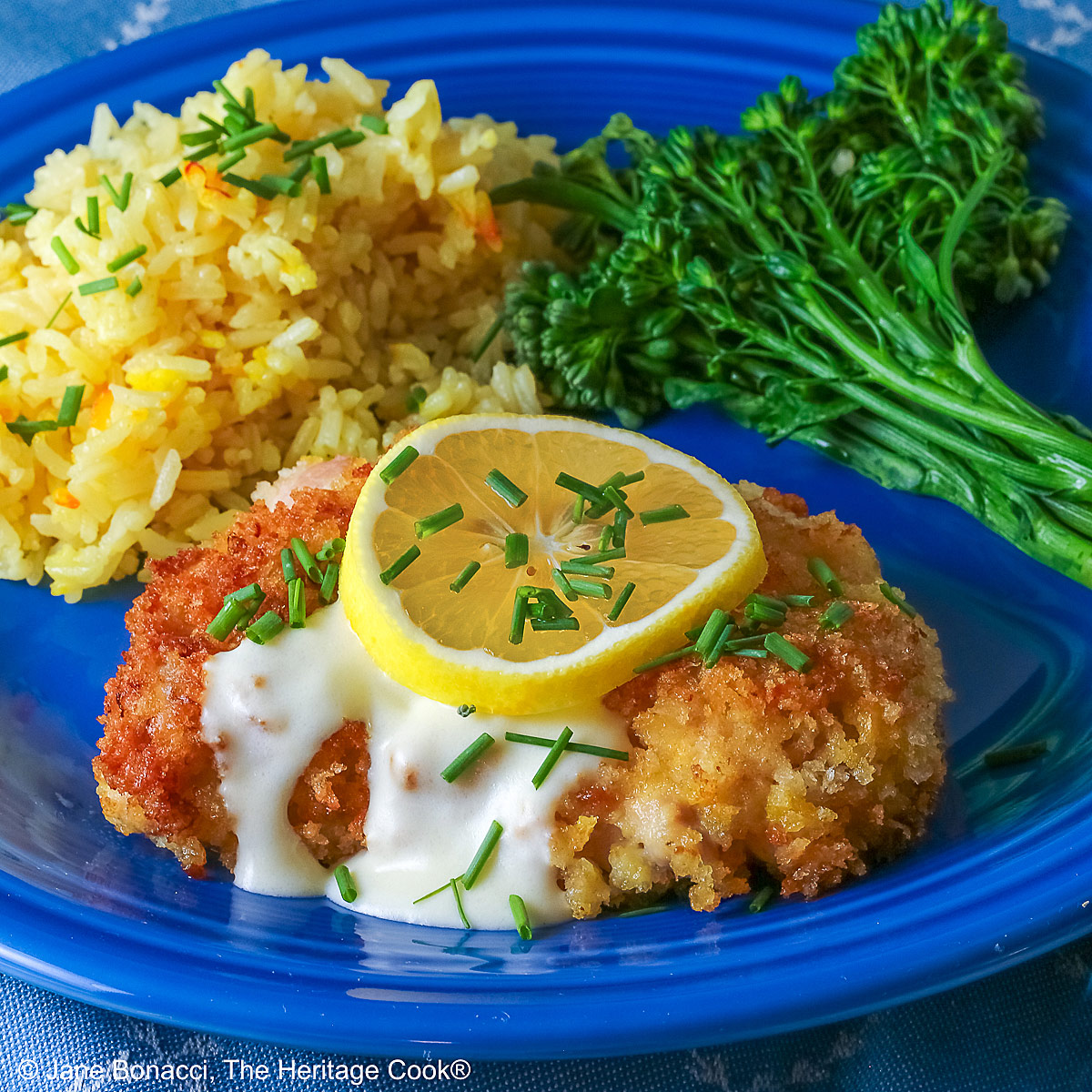 A golden brown crisp fried chicken thigh with a lemon-cream-garlic-butter sauce over the center, topped with a lemon slice and chopped chives, flanked with yellow saffron rice and green broccolini on a blue plate with a blue cloth beneath © 2026 Jane Bonacci, The Heritage Cook.