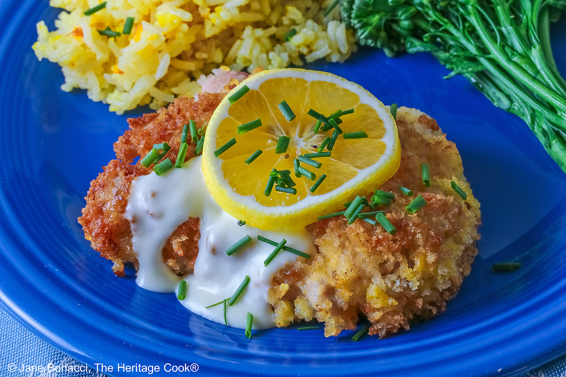 A golden brown crisp fried chicken thigh with a lemon-cream-garlic-butter sauce over the center, topped with a lemon slice and chopped chives, flanked with yellow saffron rice and green broccolini on a blue plate with a blue cloth beneath © 2026 Jane Bonacci, The Heritage Cook.