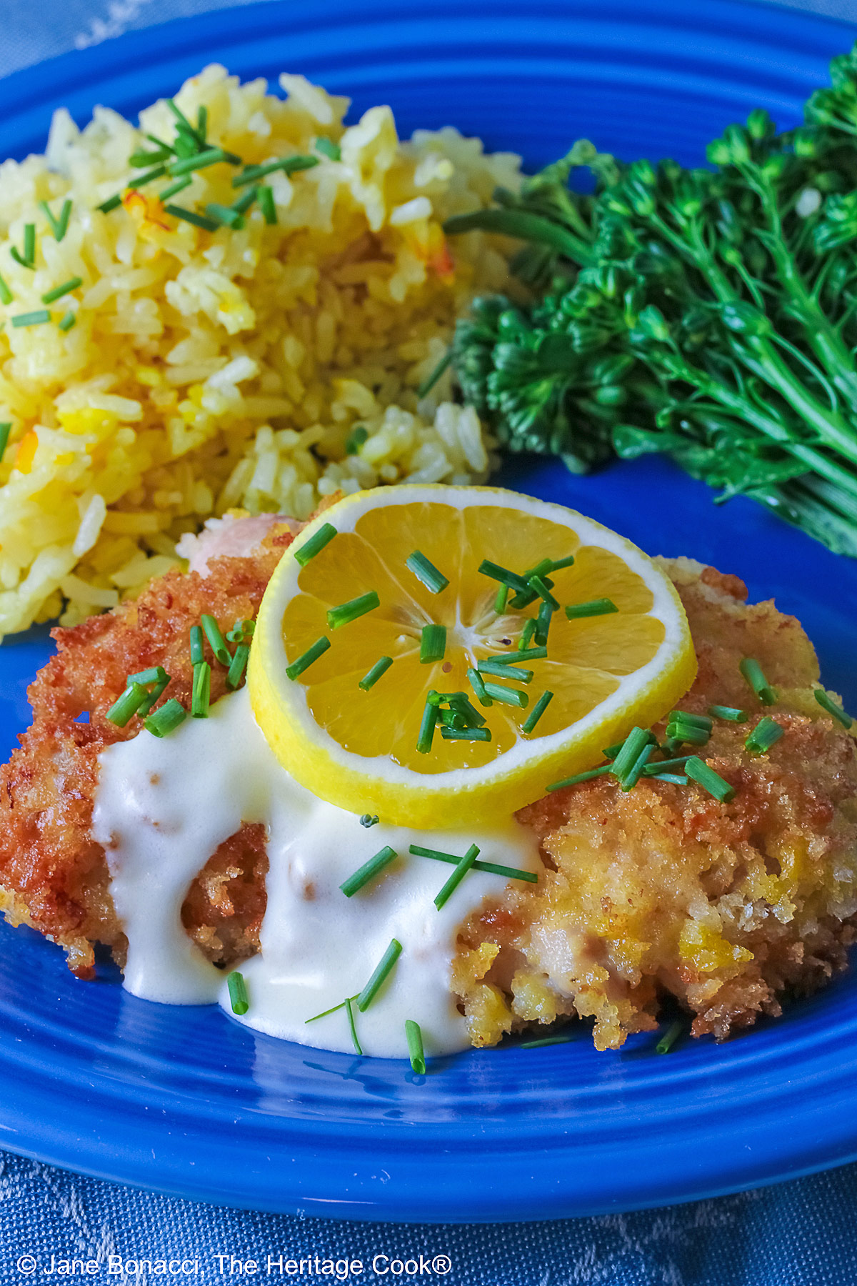 A golden brown crisp fried chicken thigh with a lemon-cream-garlic-butter sauce over the center, topped with a lemon slice and chopped chives, flanked with yellow saffron rice and green broccolini on a blue plate with a blue cloth beneath © 2026 Jane Bonacci, The Heritage Cook.