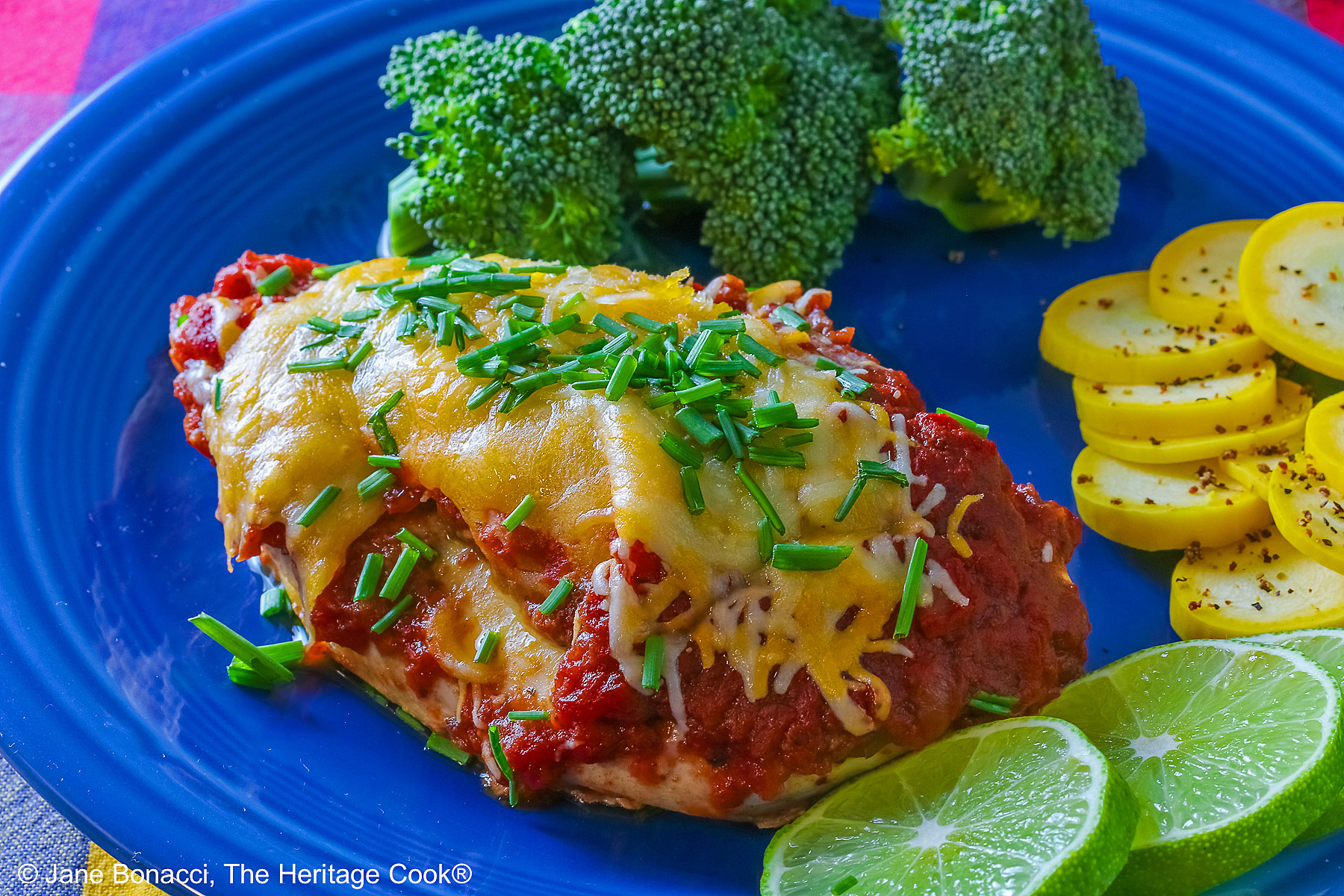 Varying angles of a salsa and melted cheese covered chicken breast with a sprinkling of chives on a blue plate surrounded by broccoli, squash coins, and lime slices, sitting on a brightly colored square cloths © 2026 Jane Bonacci, The Heritage Cook.