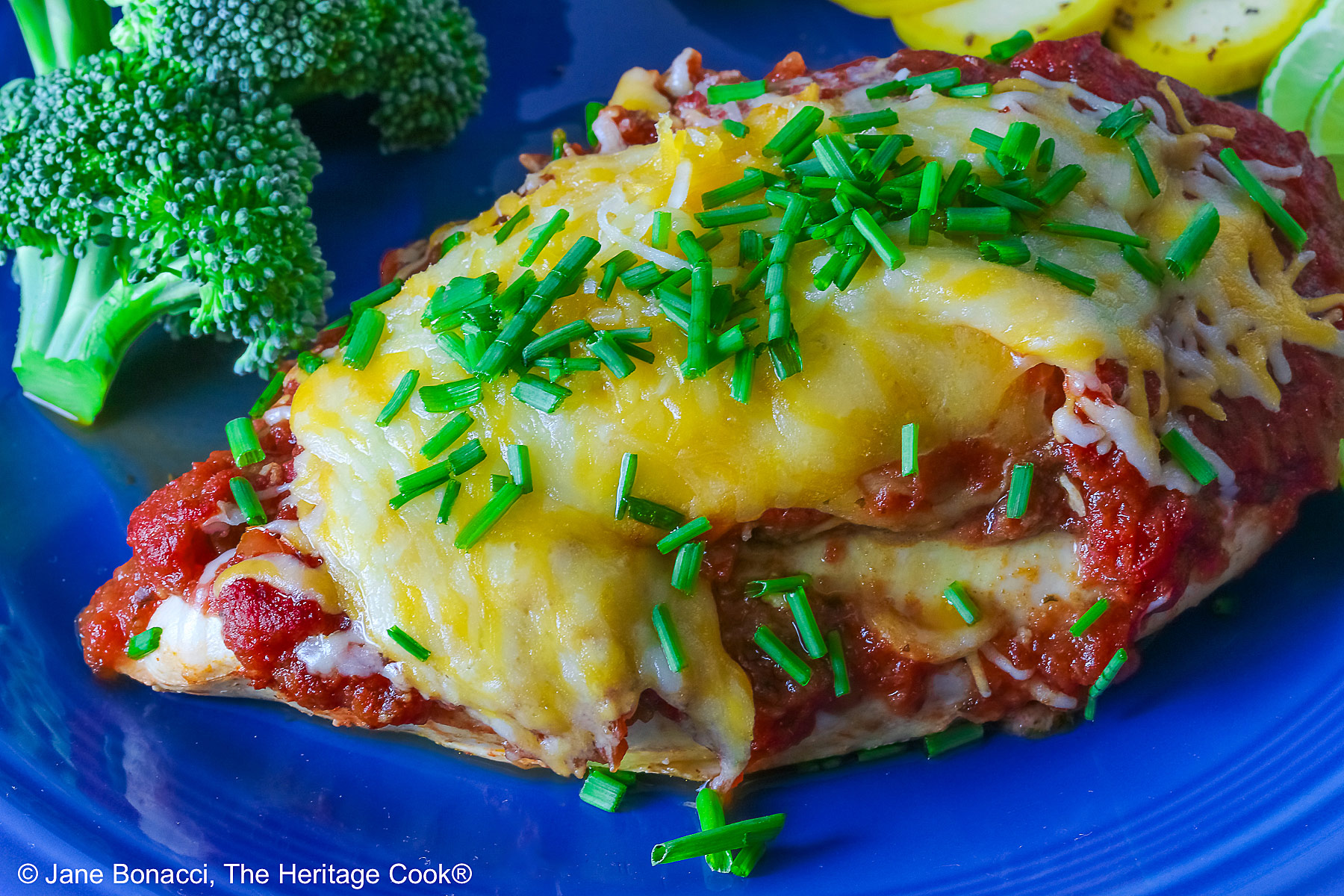 Varying angles of a salsa and melted cheese covered chicken breast with a sprinkling of chives on a blue plate surrounded by broccoli, squash coins, and lime slices, sitting on a brightly colored square cloths © 2026 Jane Bonacci, The Heritage Cook.