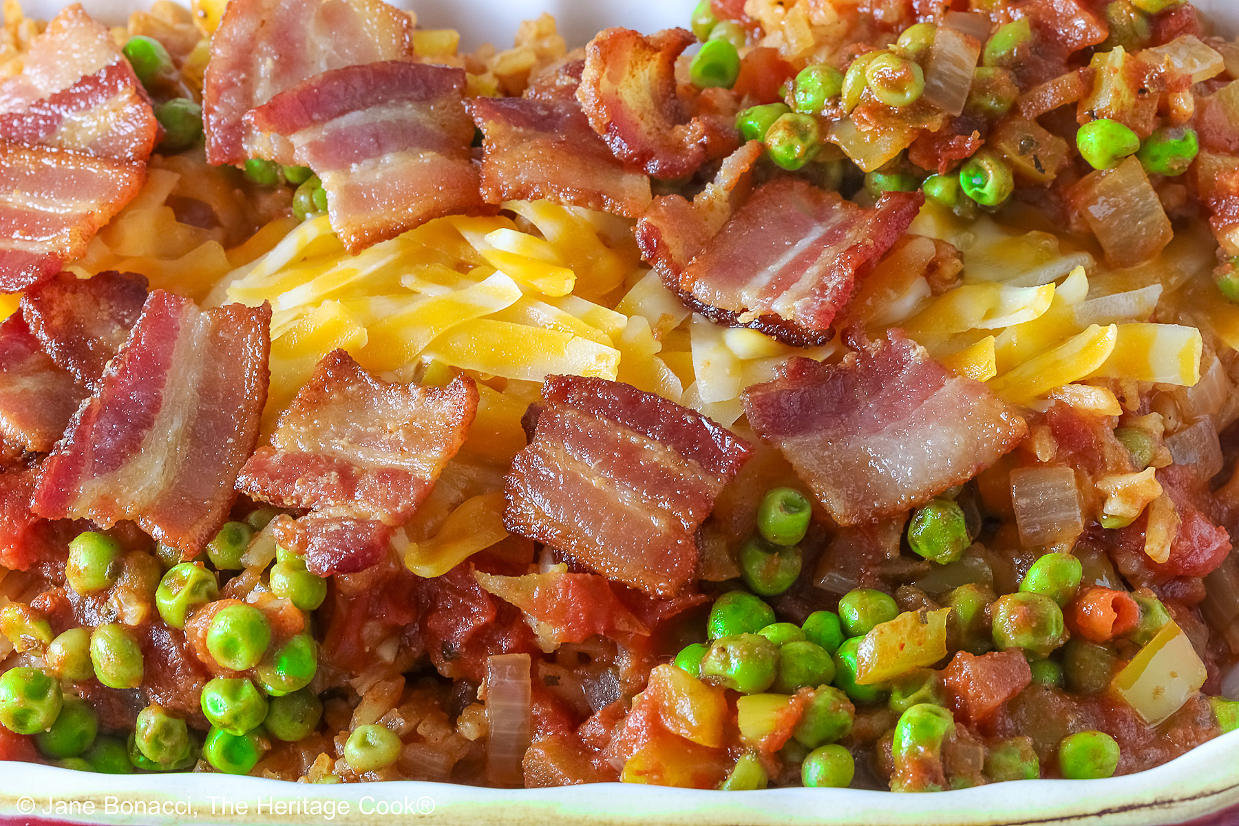 Small red rectangular casserole dish piled with Tex-Mex Rice with Bacon, with green peas scattered over the surface and shredded cheese down the center, then sprinkled with crispy bacon pieces, sitting on a blue cloth on a mottled gray background © 2026 Jane Bonacci, The Heritage Cook.