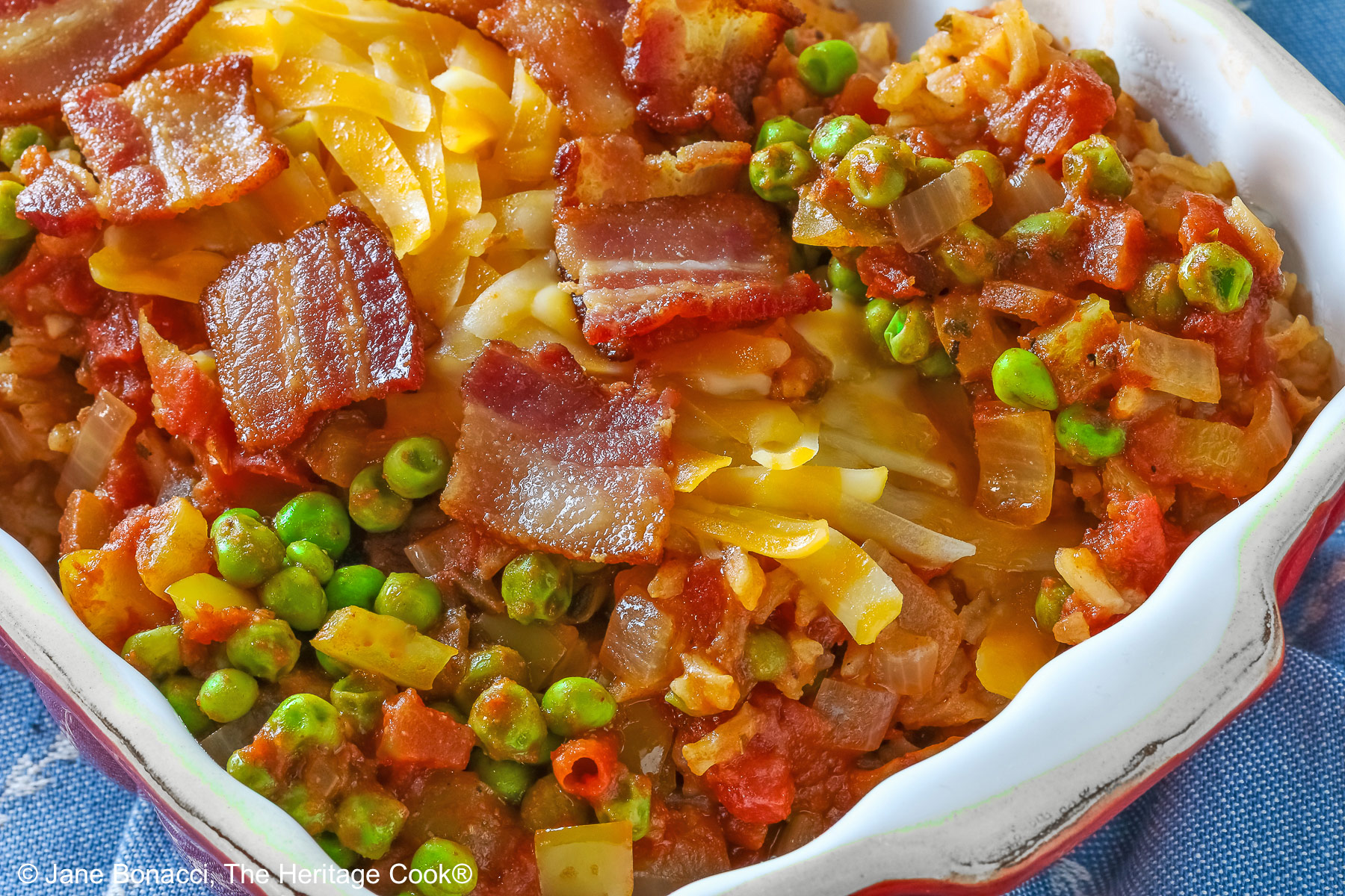 Small red rectangular casserole dish piled with Tex-Mex Rice with Bacon, with green peas scattered over the surface and shredded cheese down the center, then sprinkled with crispy bacon pieces, sitting on a blue cloth on a mottled gray background © 2026 Jane Bonacci, The Heritage Cook.
