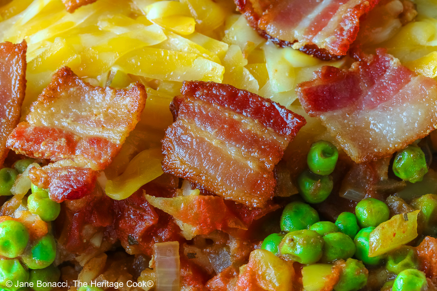 Small red rectangular casserole dish piled with Tex-Mex Rice with Bacon, with green peas scattered over the surface and shredded cheese down the center, then sprinkled with crispy bacon pieces, sitting on a blue cloth on a mottled gray background © 2026 Jane Bonacci, The Heritage Cook.