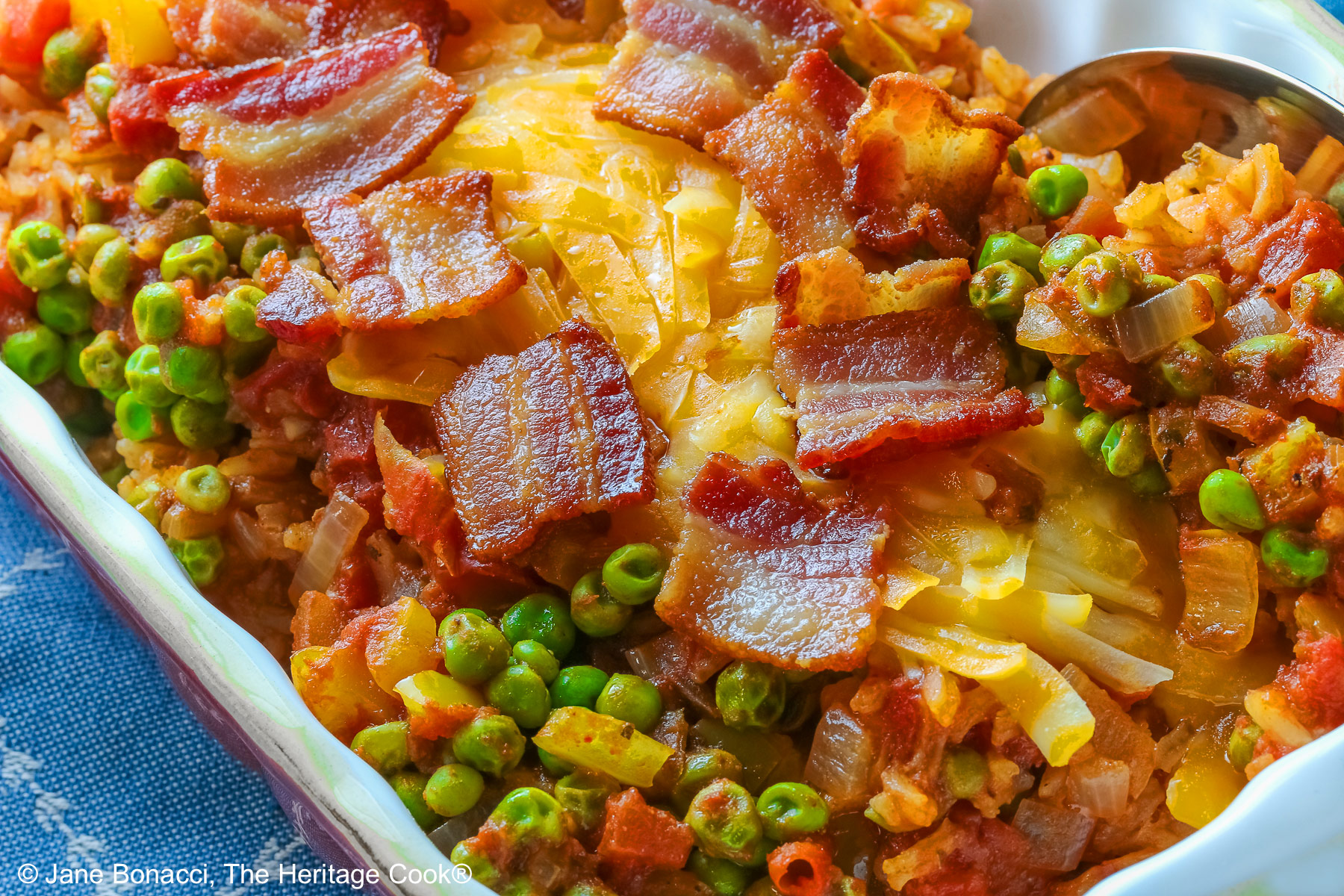 Small red rectangular casserole dish piled with Tex-Mex Rice with Bacon, with green peas scattered over the surface and shredded cheese down the center, then sprinkled with crispy bacon pieces, sitting on a blue cloth on a mottled gray background © 2026 Jane Bonacci, The Heritage Cook.