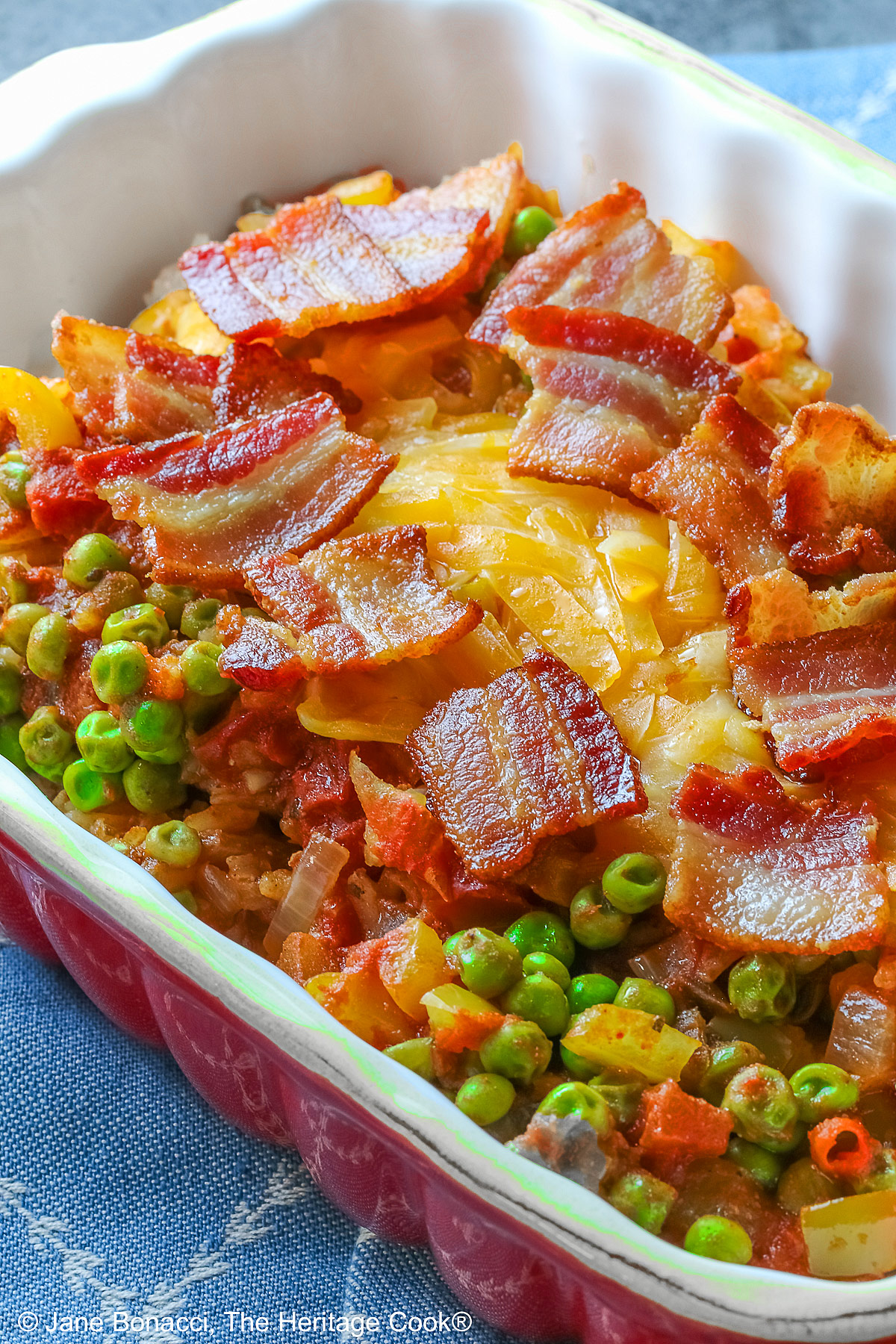 Small red rectangular casserole dish piled with Tex-Mex Rice with Bacon, with green peas scattered over the surface and shredded cheese down the center, then sprinkled with crispy bacon pieces, sitting on a blue cloth on a mottled gray background © 2026 Jane Bonacci, The Heritage Cook.