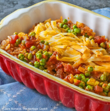 Small red rectangular casserole dish piled with Tex-Mex Rice with Bacon, with green peas scattered over the surface and shredded cheese down the center, then sprinkled with crispy bacon pieces, sitting on a blue cloth on a mottled gray background © 2026 Jane Bonacci, The Heritage Cook.
