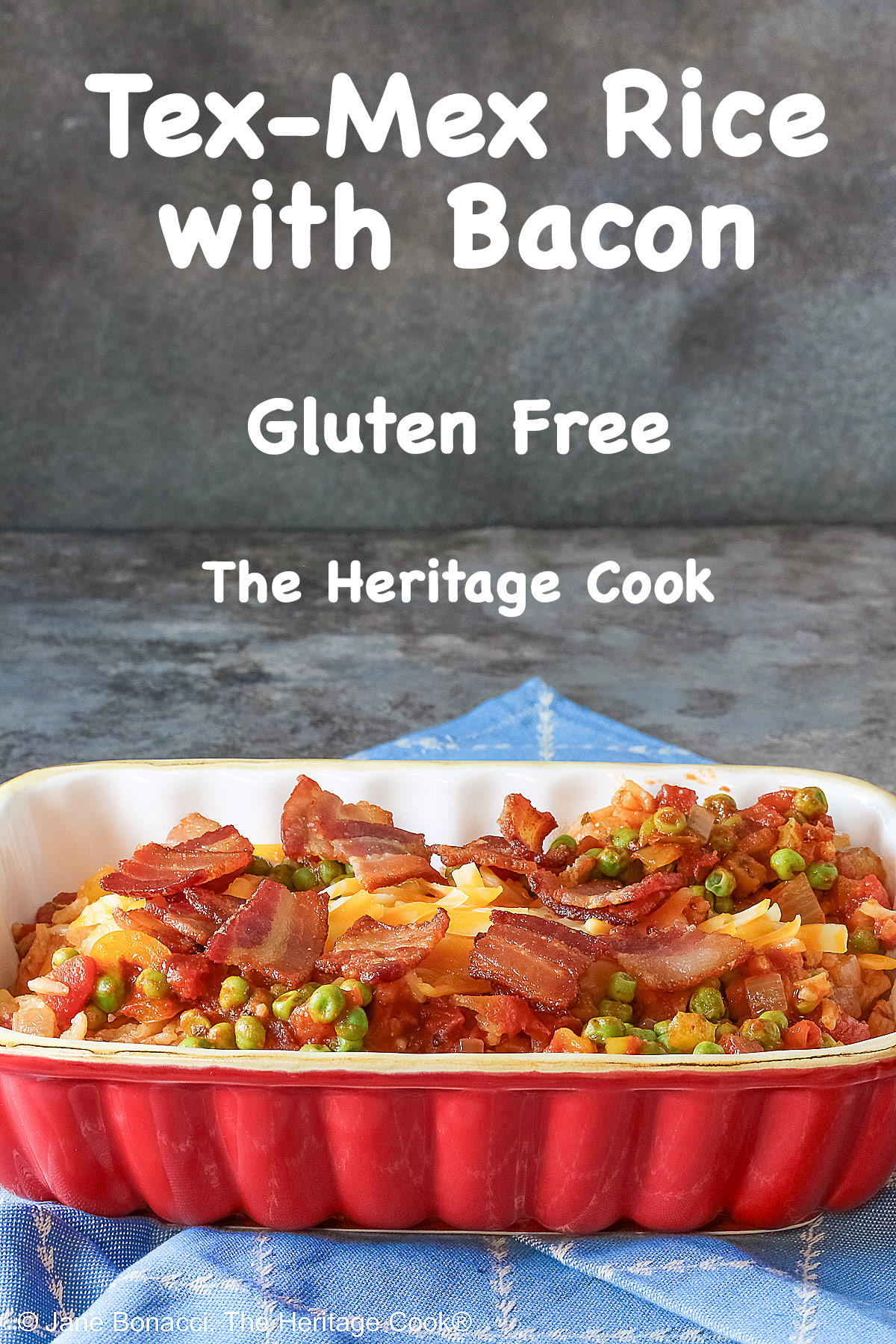 Small red rectangular casserole dish piled with Tex-Mex Rice with Bacon, with green peas scattered over the surface and shredded cheese down the center, then sprinkled with crispy bacon pieces, sitting on a blue cloth on a mottled gray background © 2026 Jane Bonacci, The Heritage Cook.