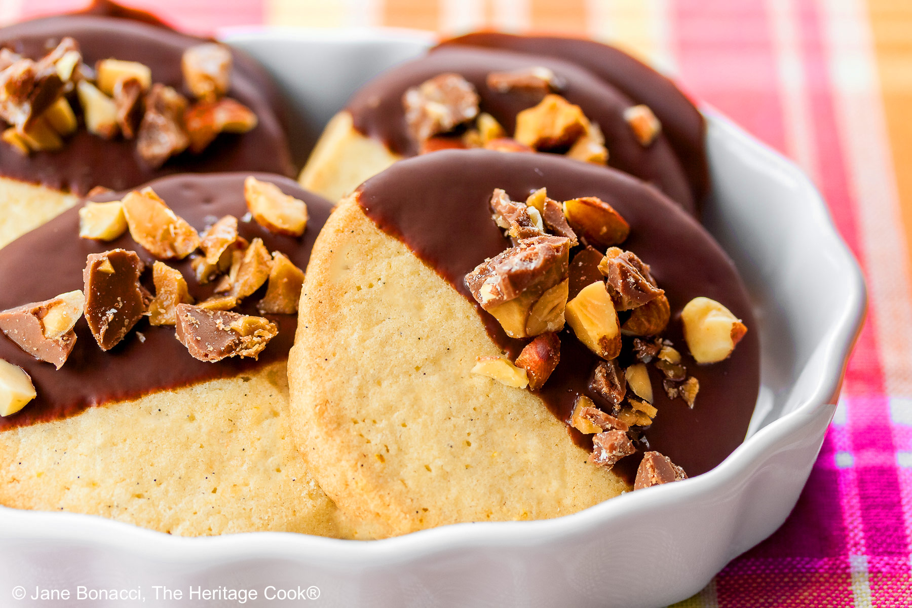 4 chocolate dipped sugar cookies topped with chopped toffee almond pieces, assembled in a white brulee dish sitting on a pink and orange plaid cloth © 2026 Jane Bonacci, The Heritage Cook.
