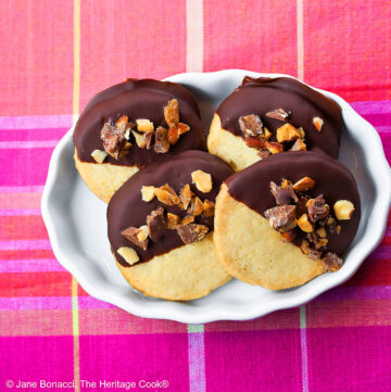 4 chocolate dipped sugar cookies topped with chopped toffee almond pieces, assembled in a white brulee dish sitting on a pink and orange plaid cloth © 2026 Jane Bonacci, The Heritage Cook.