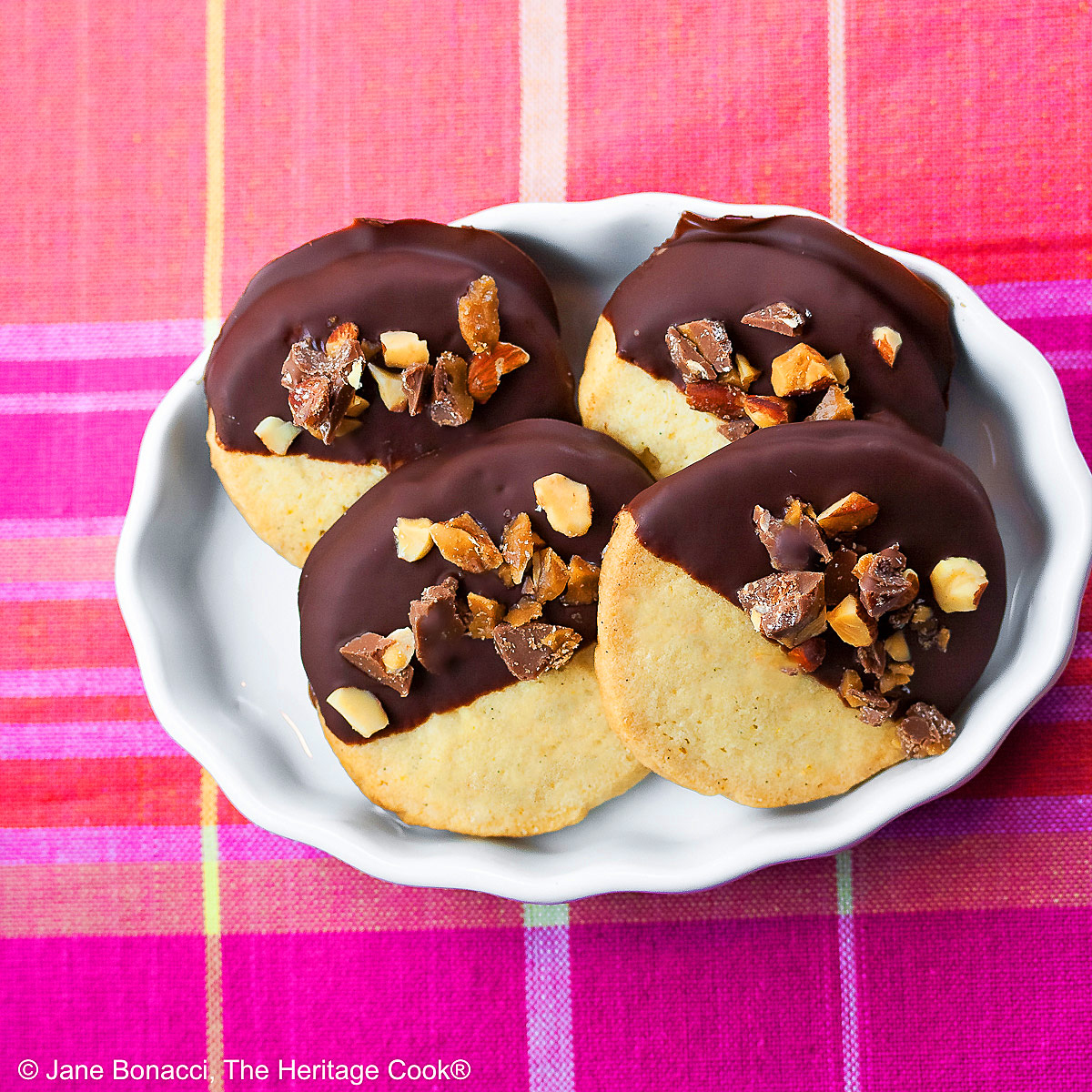 4 chocolate dipped sugar cookies topped with chopped toffee almond pieces, assembled in a white brulee dish sitting on a pink and orange plaid cloth © 2026 Jane Bonacci, The Heritage Cook.